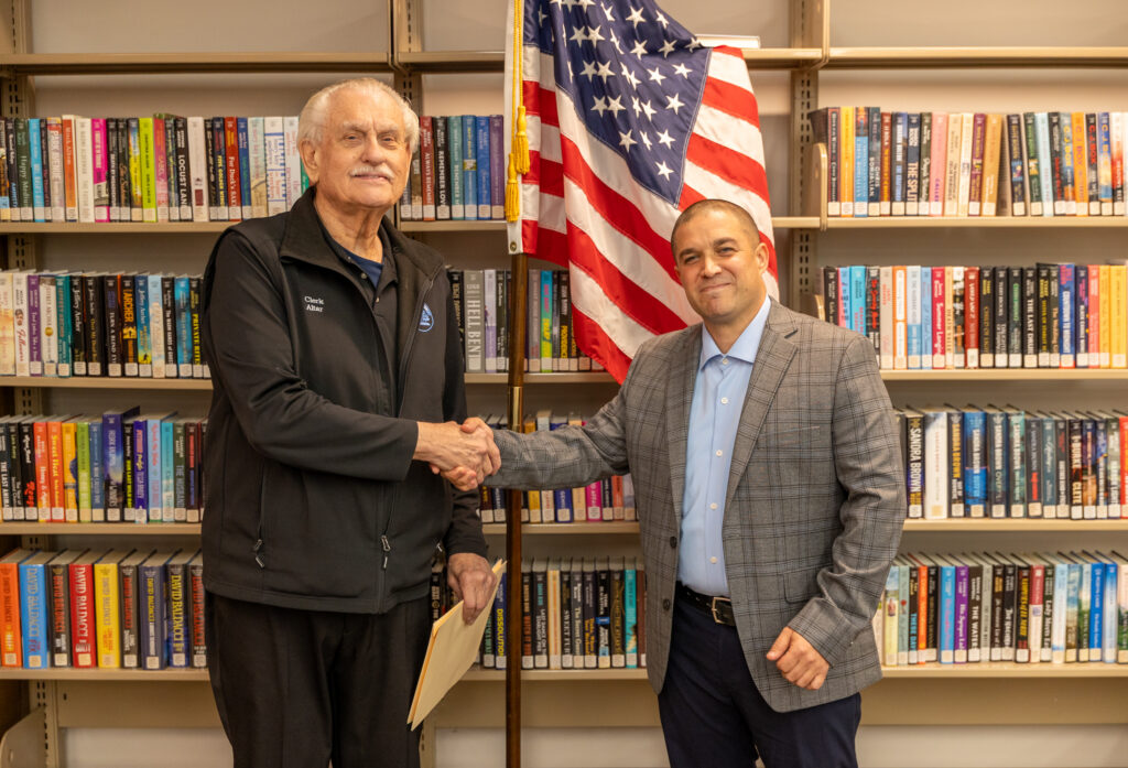 A man shakes a man's hand in front of bookshelves and an American Flag.
